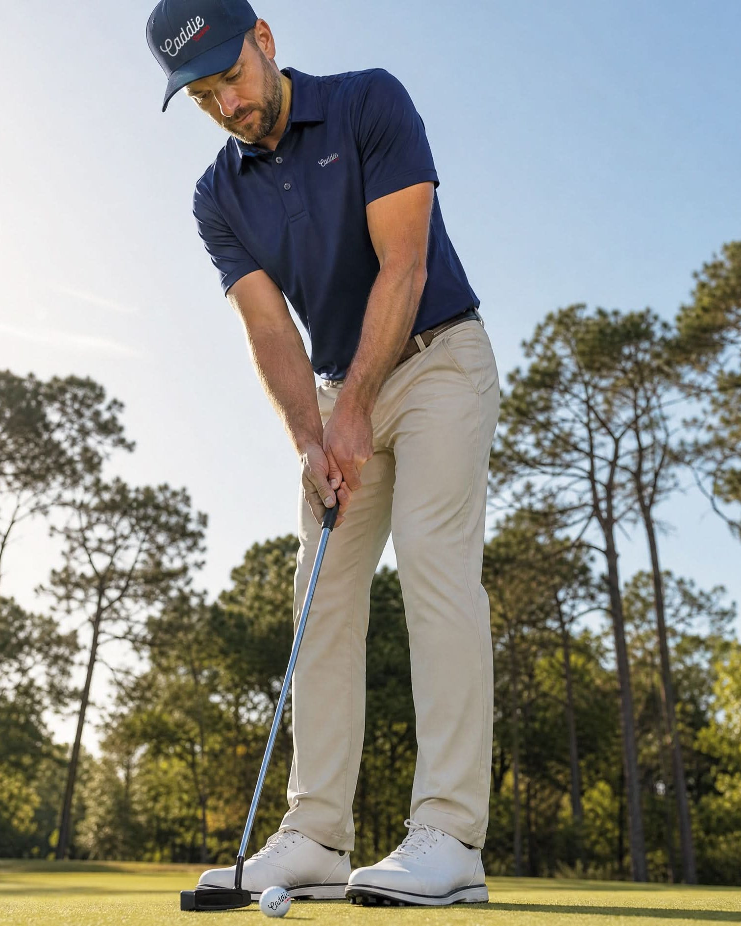A man in a navy blue polo shirt, khaki pants, white golf shoes, and a navy Caddie Couture cap is shown from the waist down on a golf course, preparing to putt with a putter.