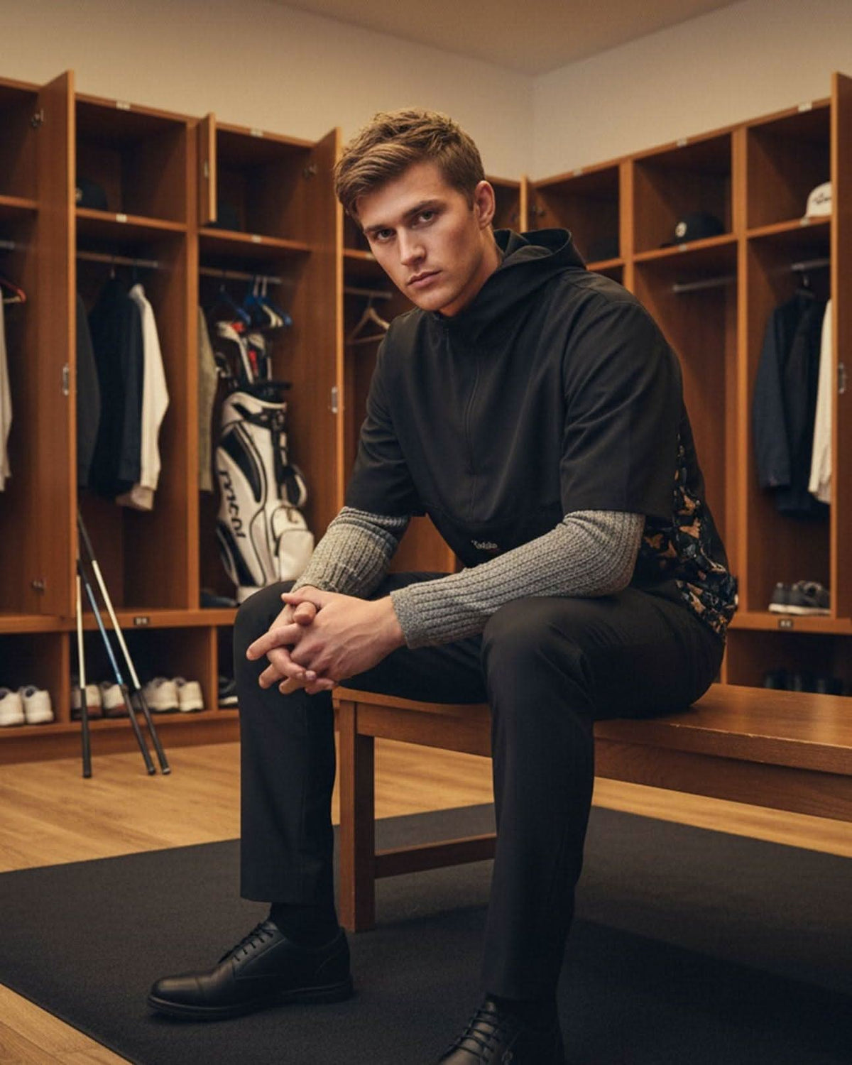 Man sitting in a locker room wearing a black windbreaker and gray sweater.