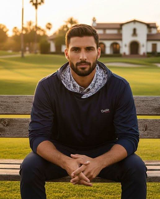 Man sitting on a wooden bench in a park with a building and trees in the background wearing desert golf hoodie