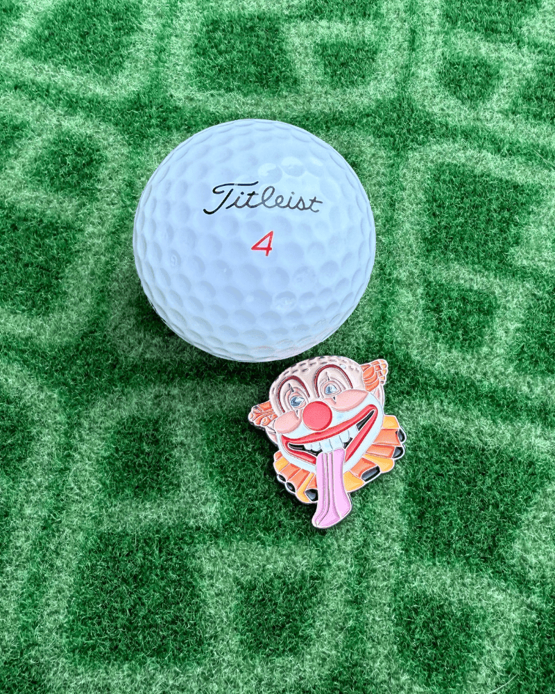 Golf ball with 'Titleist' branding on a green textured surface with Clown Golf Ball Marker Next to it
