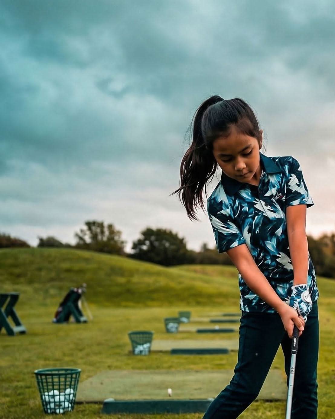 Young girl playing golf on a grassy course with trees and a cloudy sky in the background.
