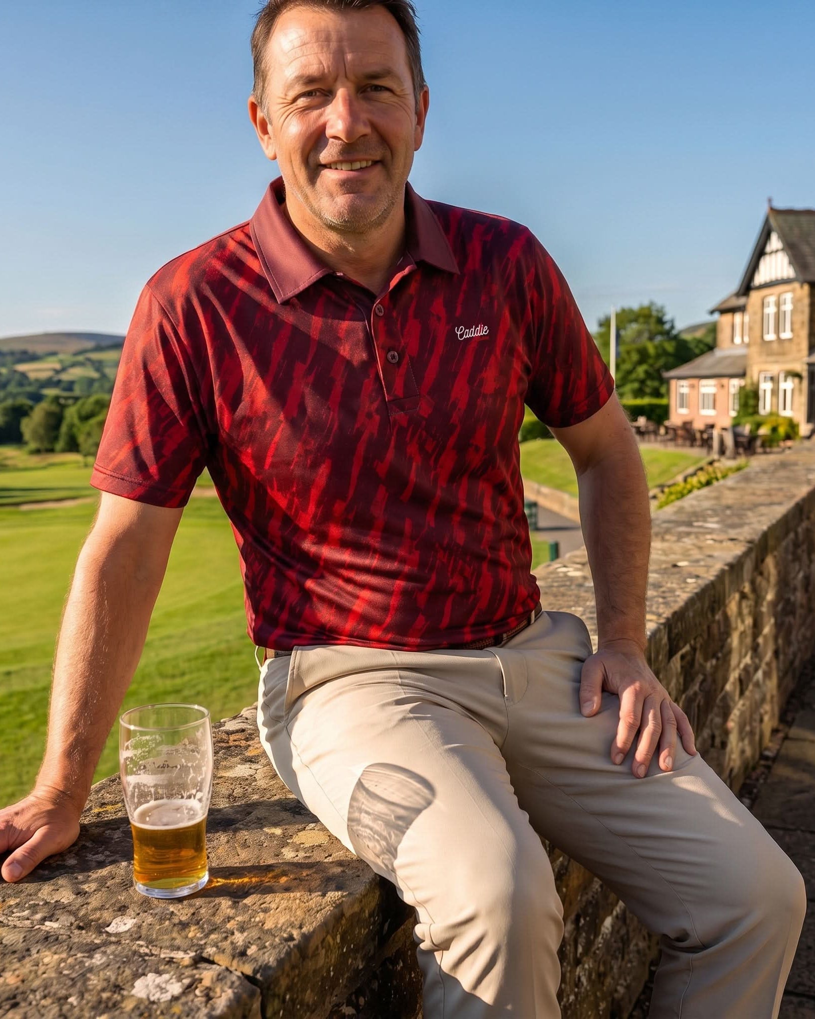 A smiling man wearing a red and black patterned polo shirt and beige trousers sits on a stone wall outdoors, with a glass of beer in front of him and a country estate in the background.