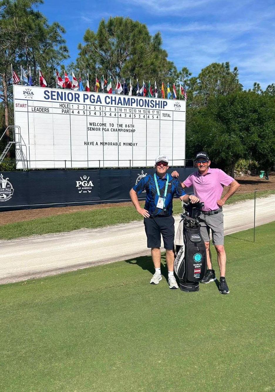 Two golfers posing with a golf bag in front of a large scoreboard at the Senior PGA Championship.