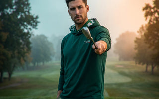 man pointing golf club at the camera wearing green golf hoodie on a golf course