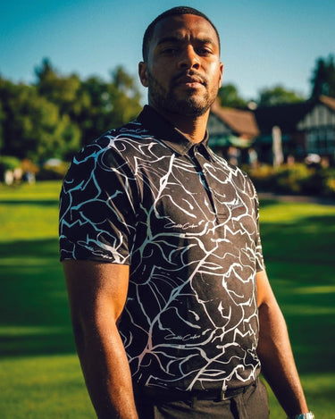 Man wearing a patterned shirt standing outdoors with greenery in the background
