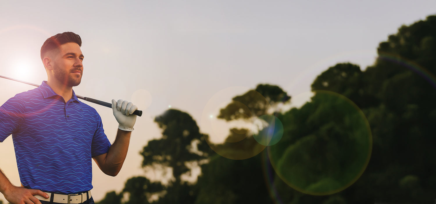 Man holding a golf club with trees in the background wearing blue waves golf polo