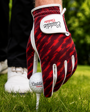 A close-up of a person's hand wearing a red and black patterned Caddie golf glove, holding a golf ball on a tee over a grassy golf course.