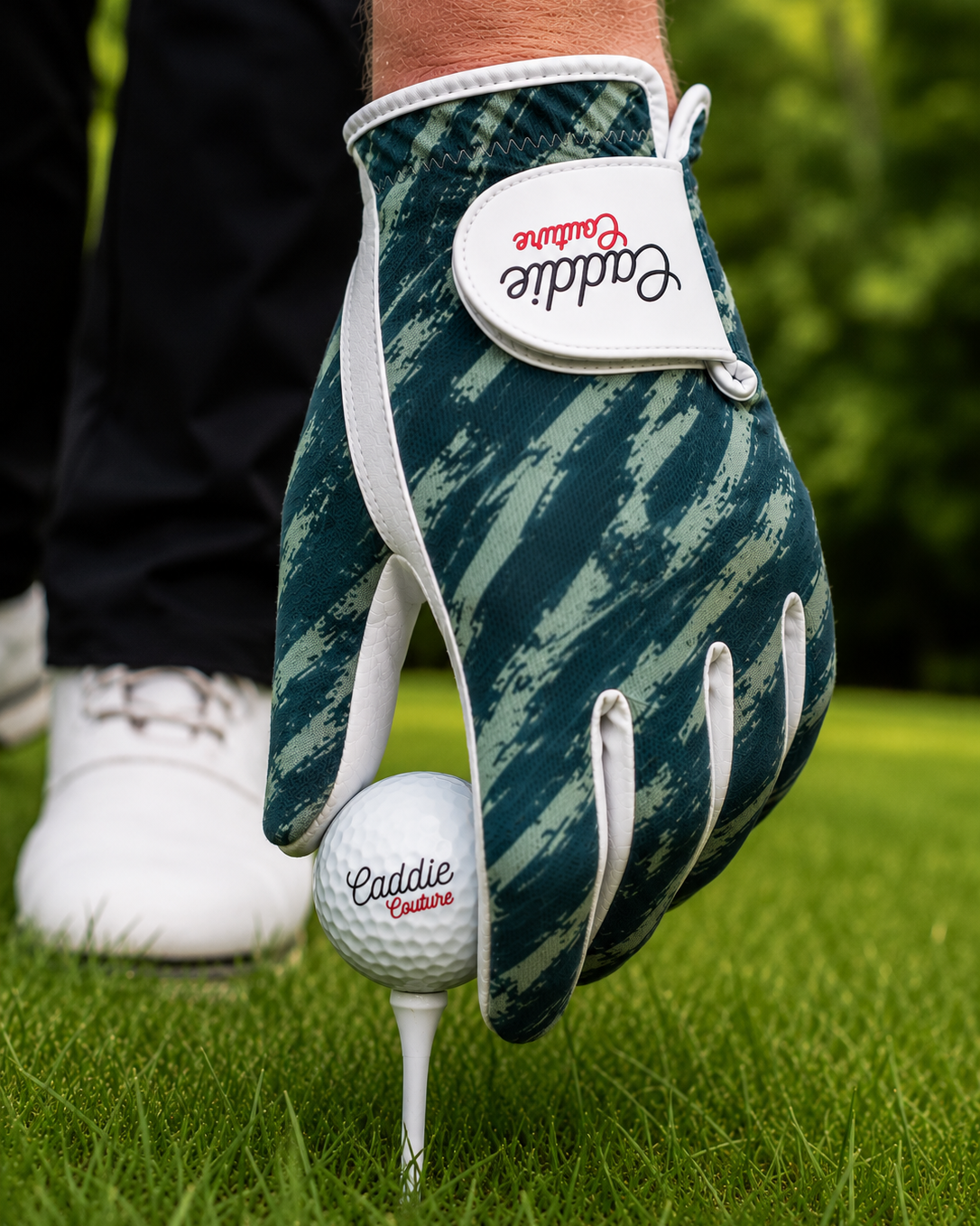 A close-up of a golf glove worn on a hand, holding a golf ball on a tee. The glove has a dark teal and green camouflage pattern with white trim and a white brand patch that reads 'Caddie Collection'.
