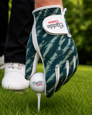 A close-up of a golf glove worn on a hand, holding a golf ball on a tee. The glove has a dark teal and green camouflage pattern with white trim and a white brand patch that reads 'Caddie Collection'.