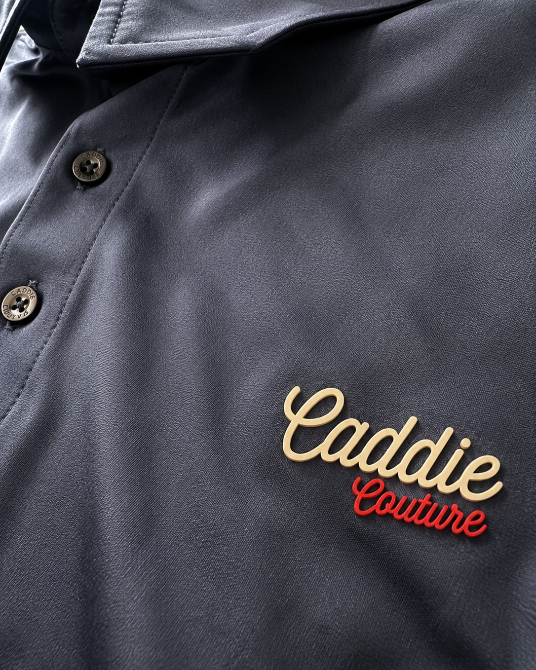 Close-up of a dark charcoal gray button-up shirt with a collar, featuring an embroidered logo on the chest that reads 'Caddie Couture' in gold and red.