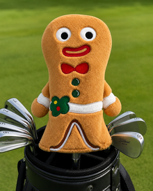 A plush gingerbread person golf club headcover sits on top of a black golf bag, with metal golf clubs visible in the bag and a blurred green grass background behind it.