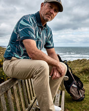 A man sitting on a wooden bench outdoors by the ocean, wearing a teal and light blue patterned polo shirt, khaki pants, and a baseball cap, with a golf bag beside him.