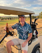 A smiling man sitting in a beige golf cart on a golf course at sunset, holding a beer bottle. He is wearing a blue and white tropical print polo shirt, beige shorts, and a baseball cap.