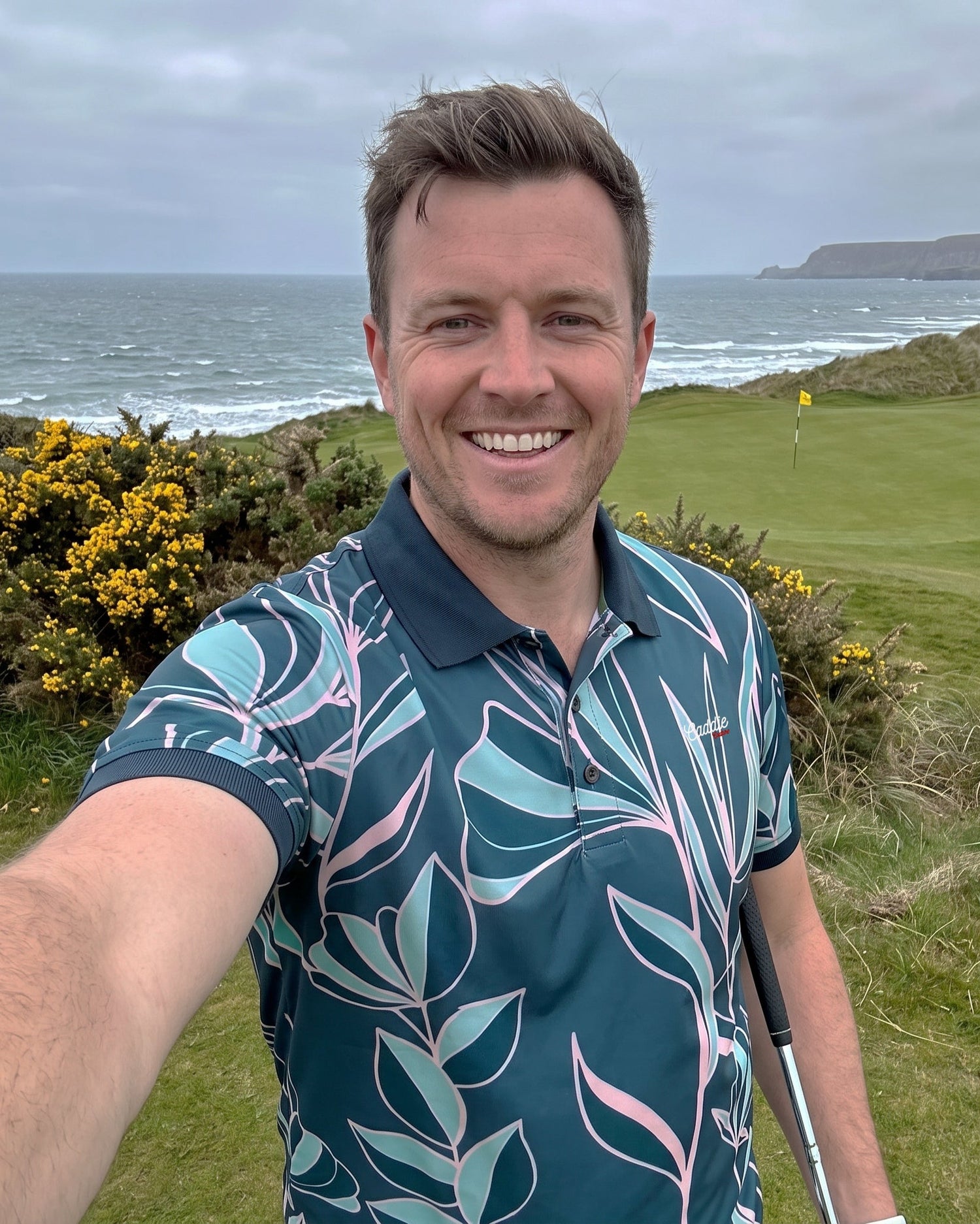 A smiling man takes a selfie outdoors on a golf course, wearing a teal short-sleeve polo shirt with a light blue and pink tropical leaf pattern. The background shows a green grassy course, yellow gorse bushes, and the ocean under an overcast sky.