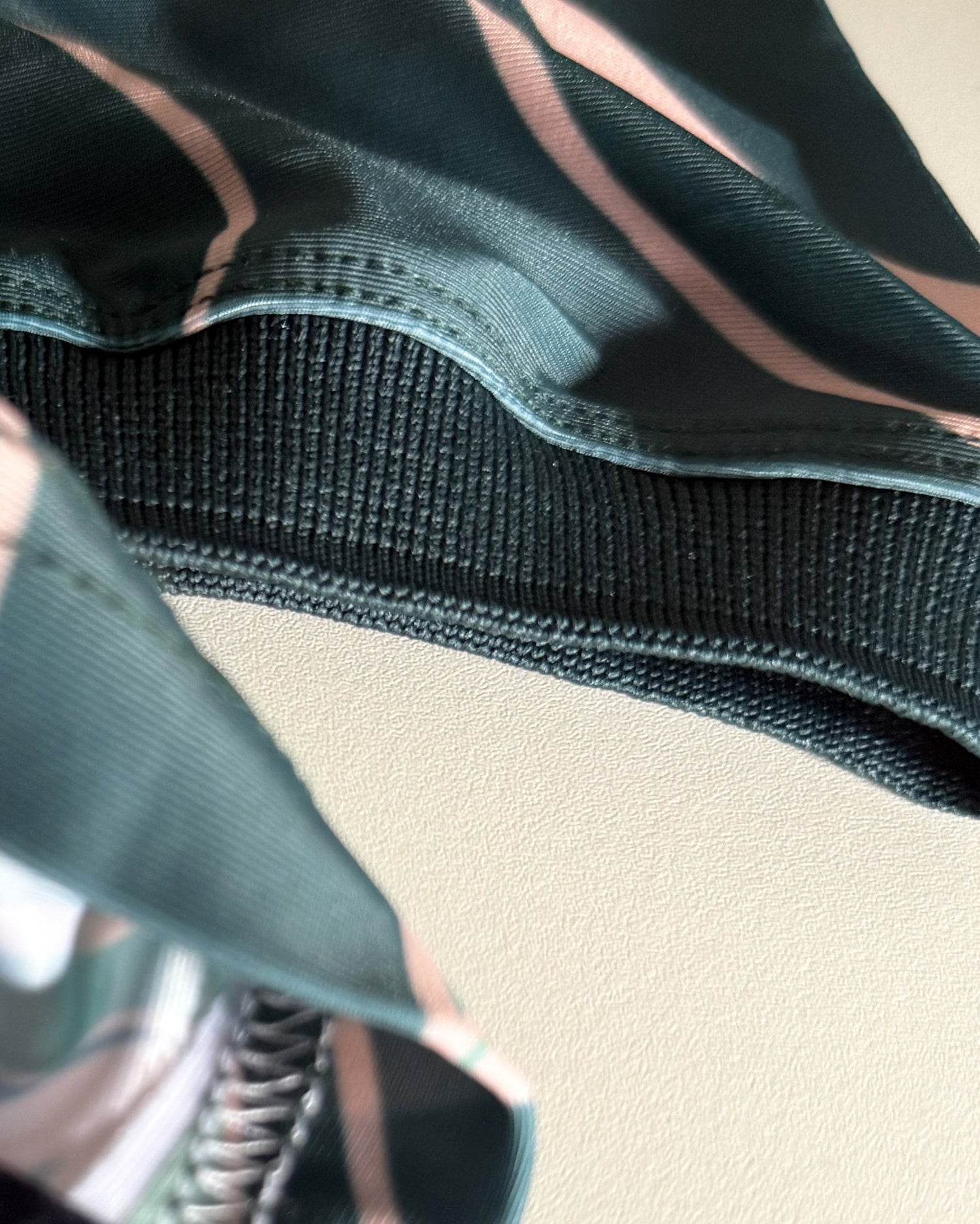 Close-up detail of a garment's interior waistband, showing a dark green elastic band with thin pink stripes and a matching striped fabric panel on a light beige background.