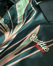 Close-up of a satin-like collared shirt with a dark green base, pale pink and sage green abstract leaf-like stripes, and embroidered 'Caddie Boutique' text on the chest.