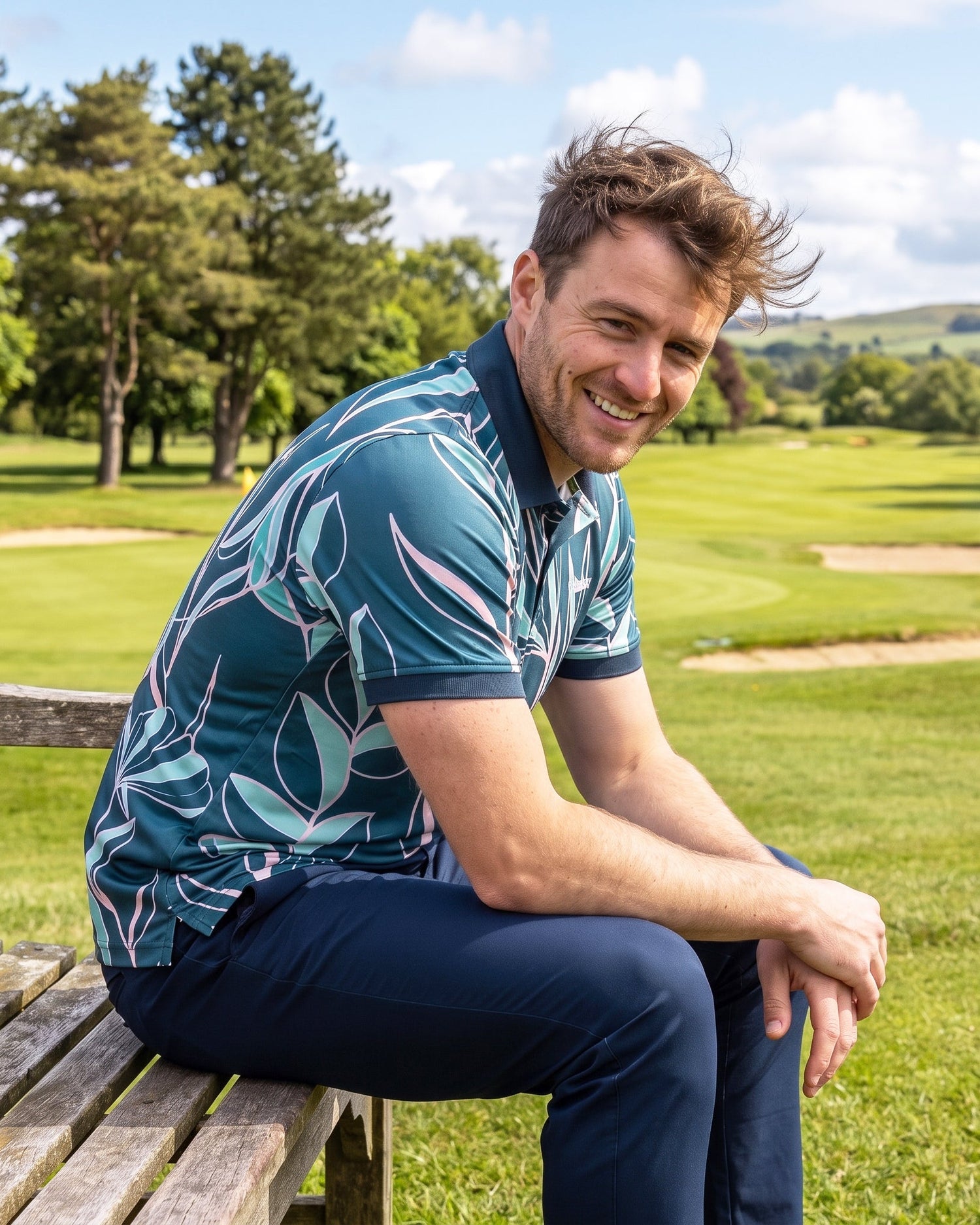 A man sitting on a wooden bench at a golf course, wearing a teal short-sleeve polo shirt with a white and light blue tropical leaf pattern and navy trousers.