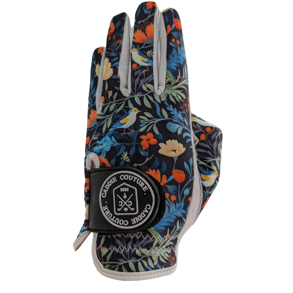 A floral patterned golf glove with multicolored flowers and green leaves on a black background, featuring a white brand label on the wrist.