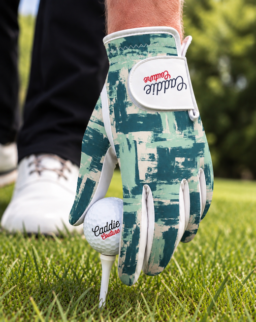 A close-up of a golf glove with a green and white brushstroke pattern, holding a golf ball on a tee on a grassy golf course. The glove features the brand name 'Caddie Outfitter' on the back and wrist strap.