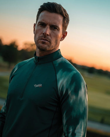 Man wearing a green jacket with a brand logo, standing in a field at sunset.