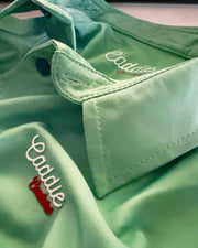 A close-up of a mint green collared shirt with 'Caddle' embroidered in white script and a small red accent on the chest, showing the collar and button details.