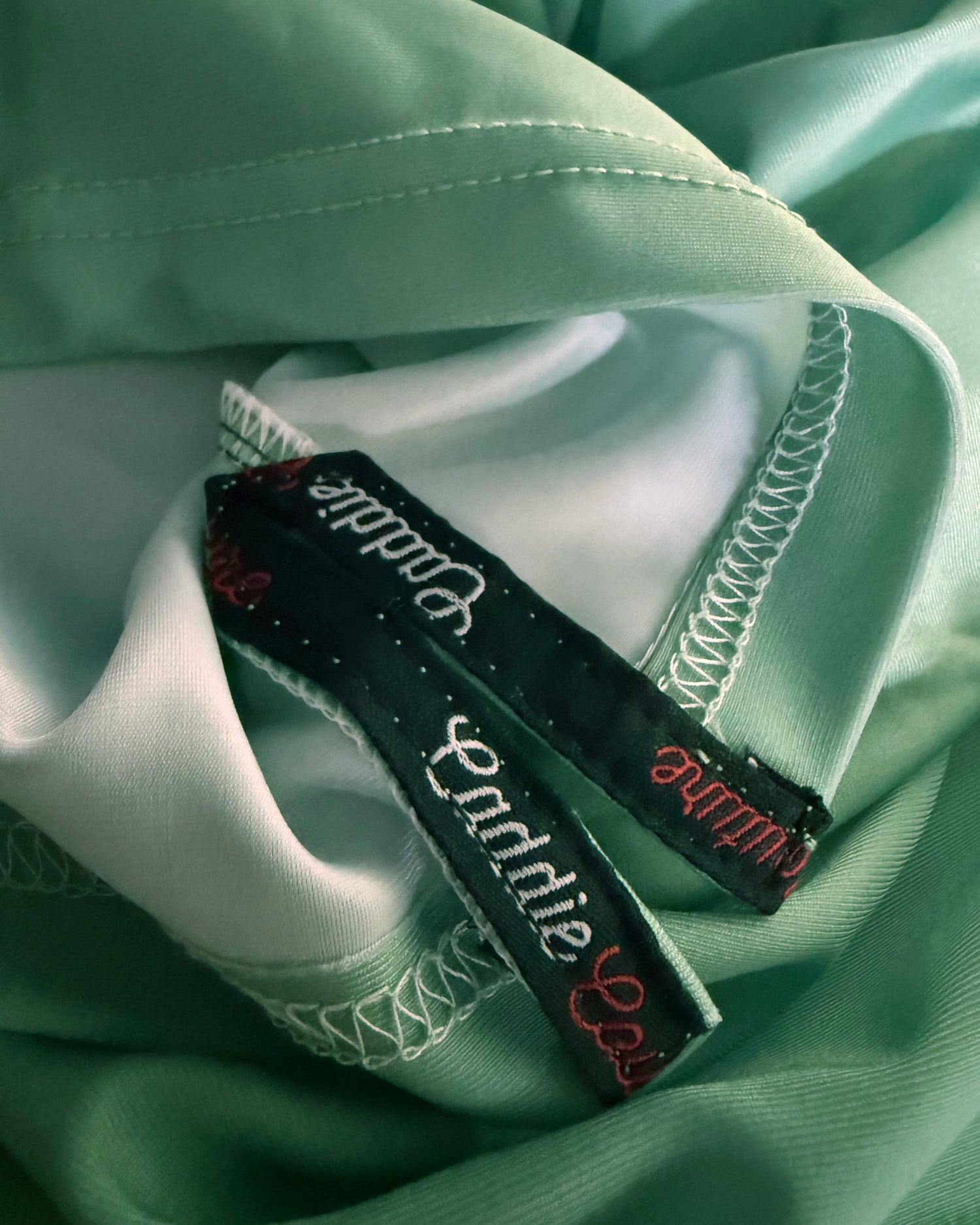Close-up of a mint green satin garment with white contrast stitching and two black fabric labels reading 'Bandle' in white script and 'Londine' in red script.