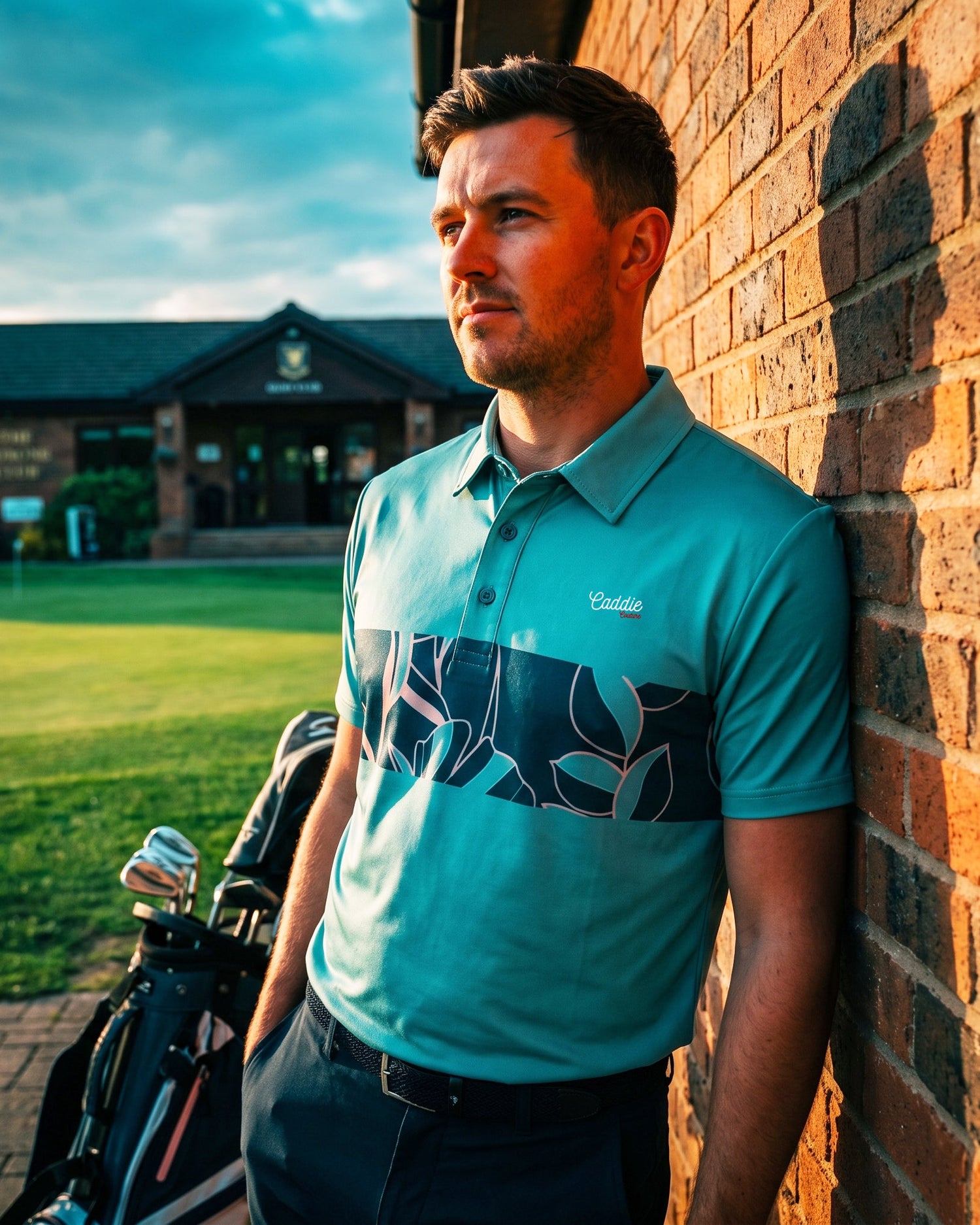 A man wearing a teal and navy patterned polo shirt leans against a brick wall outdoors, with a golf bag and a golf course building in the background.