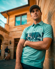 A man wearing a teal and navy patterned polo shirt and a gray cap leans against a stone wall in an outdoor courtyard setting.