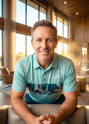 A smiling man wearing a light blue polo shirt with a dark tropical leaf pattern sits in a leather armchair in a modern, sunlit living room with wood-paneled walls and large windows.
