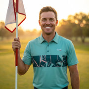 A smiling man on a golf course holds a flag while wearing a teal polo shirt with a dark teal and pink tropical leaf pattern across the chest. The background shows a warm, golden sunset over a blurred golf course.