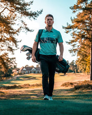 A man on a golf course wearing a teal and black polo shirt with a large 'NRA' logo, carrying a black golf bag over his shoulder.