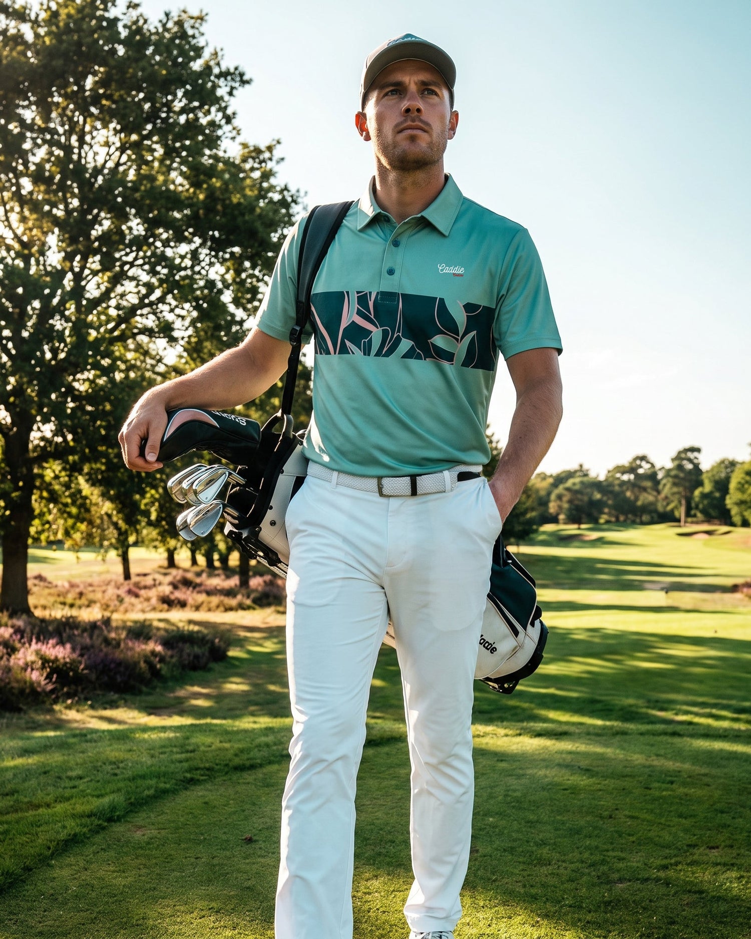 A man stands on a sunny golf course wearing a teal and black patterned polo shirt, white trousers, and a cap, while holding a golf bag over his shoulder.