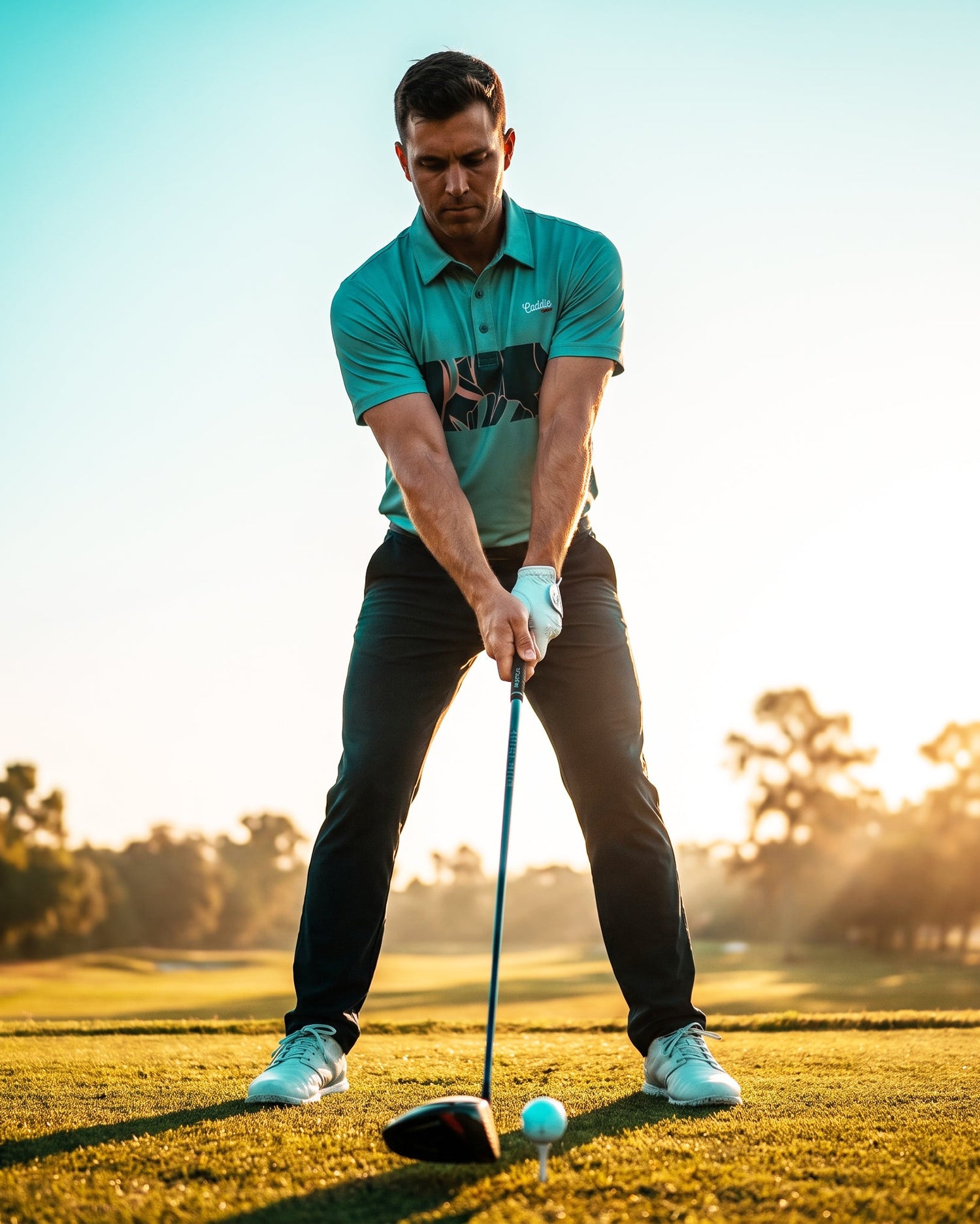 A man in a teal polo shirt and black pants is mid-swing on a golf course, with a golf club and ball in front of him. The background shows a bright, sunlit sky and blurred trees.