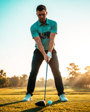 A man in a teal polo shirt and black pants is mid-swing on a golf course, with a golf club and ball in front of him. The background shows a bright, sunlit sky and blurred trees.