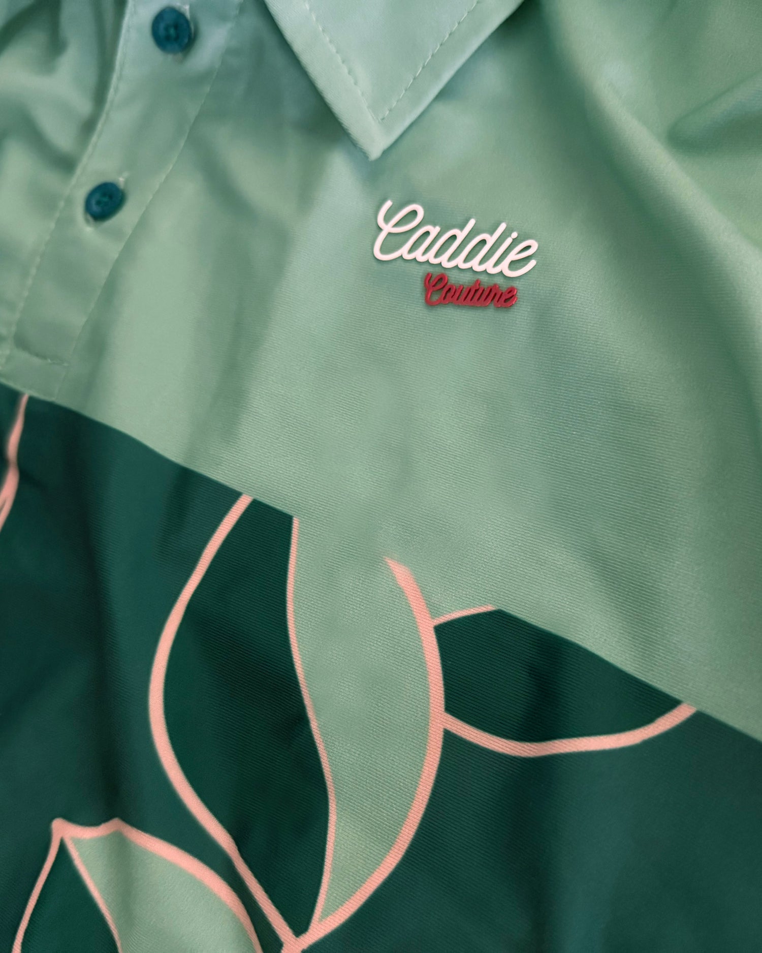 Close-up of a collared button-up shirt with a mint green upper section and a dark green tropical leaf print lower section, featuring 'Caddie Casino' embroidered on the chest.