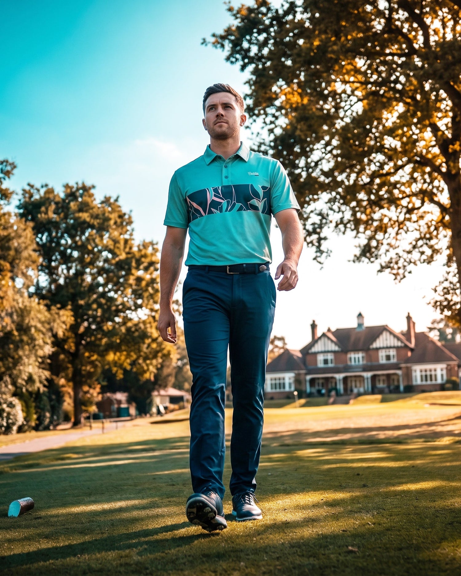 A man walking on a sunny golf course wearing a turquoise and black patterned polo shirt with dark navy trousers.