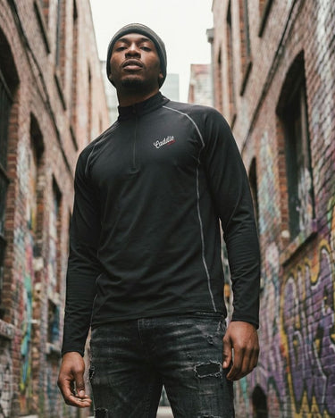 Man wearing a black long-sleeve shirt with a brand logo in an alley with graffiti.
