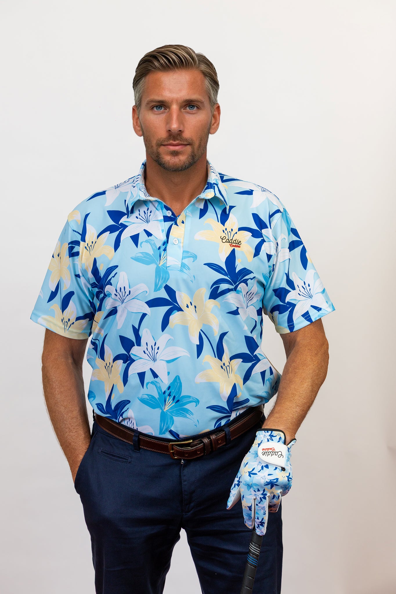 Man wearing a blue floral golf shirt and dark pants, holding a golf club with a white background
