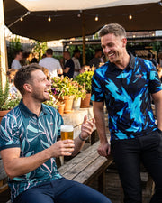 Two men laughing at an outdoor pub table, wearing matching blue and teal tropical print polo shirts. One man is seated holding a pint of beer, and the other stands beside him under a patio canopy.