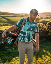 A man walking on a grassy golf course at sunset, wearing a green and teal patterned polo shirt, khaki pants, and a dark cap with a visible 'Caddie' logo. He is carrying a golf bag over his shoulder.