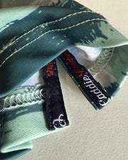 Close-up of a folded camouflage fabric item, likely a bandana or headwrap, showing a green and gray camo pattern, white contrast stitching, and two black brand tags reading 'Middle Eastern'.