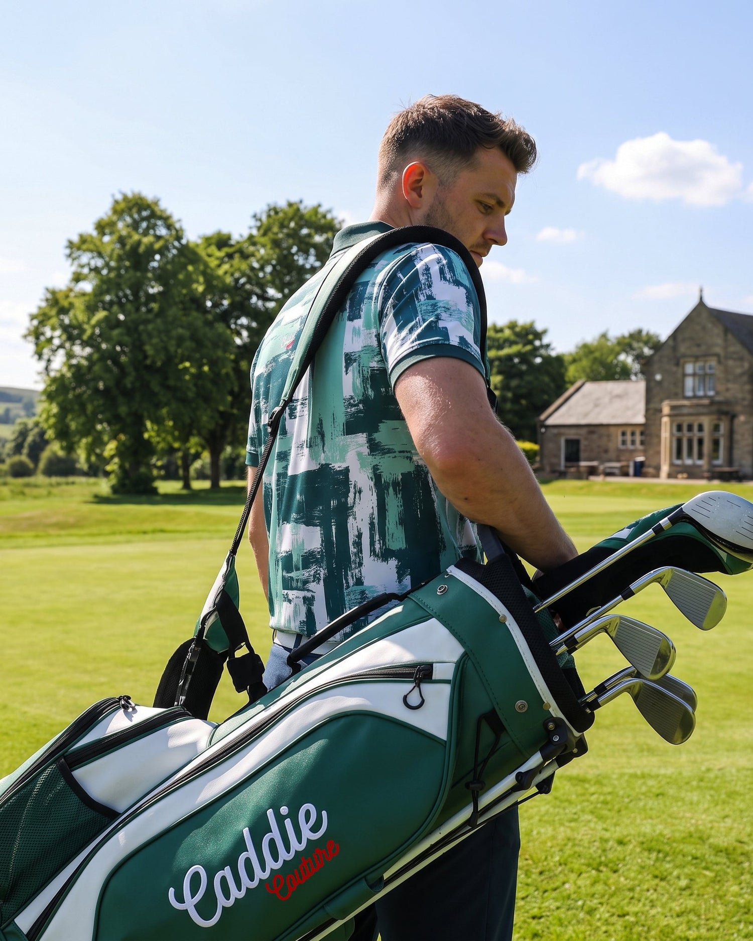 A man on a golf course carries a green and white Caddie Classic golf bag with clubs, with a stone building and trees in the background.