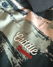 Close-up of a camouflage-patterned polo shirt with a dark gray ribbed collar and embroidered 'Caddie Boutique' logo on the chest.