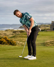 A man in a green and blue patterned polo shirt, black trousers, and white golf shoes is bent over on a golf course, holding a putter. The background features a grassy fairway, ocean coastline, and a stone building under a cloudy sky.