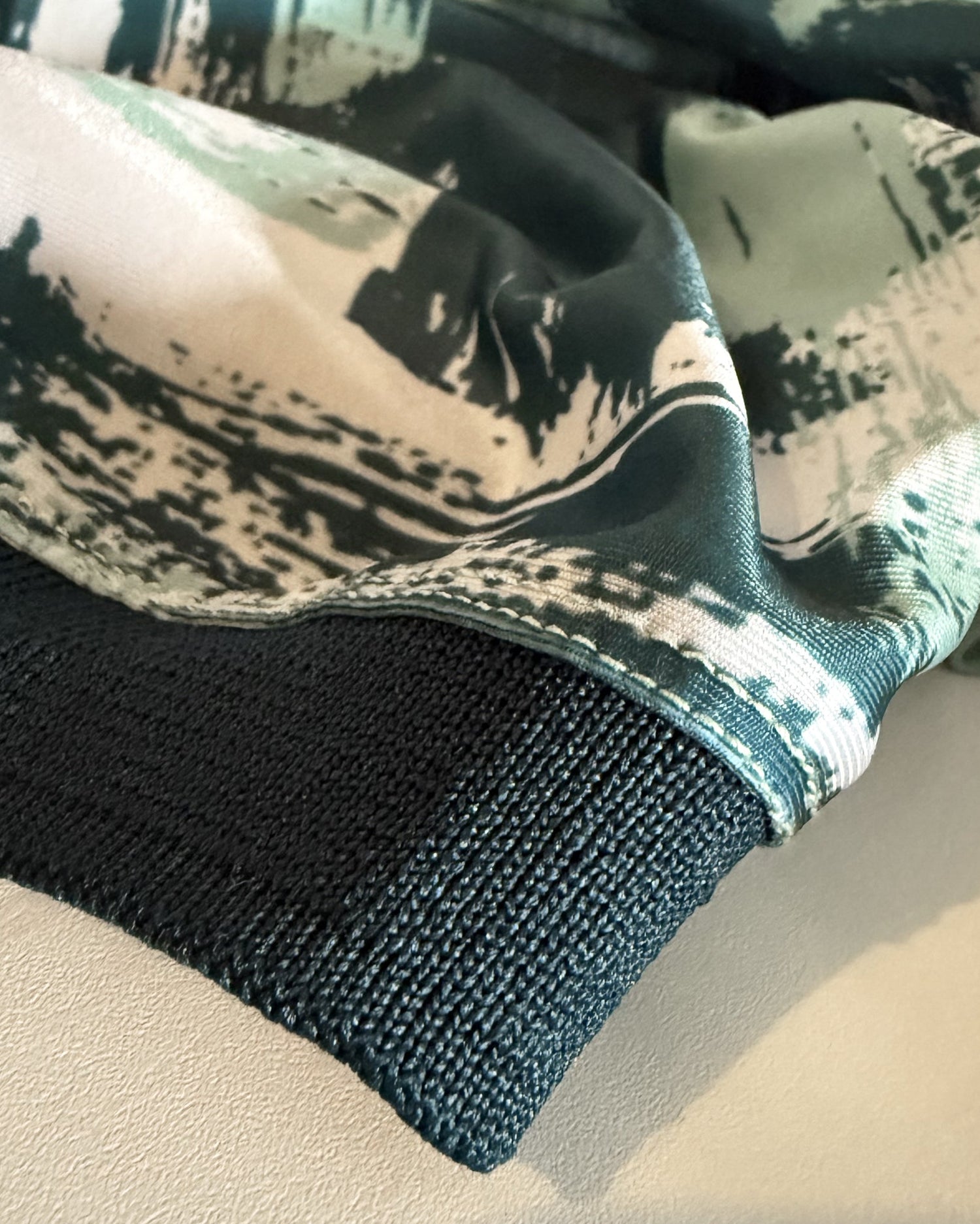 Close-up of a knit garment cuff with a dark teal ribbed edge and a matching printed fabric section above featuring a black, white, and muted green abstract brushstroke pattern.