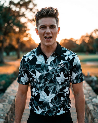 Man wearing a floral patterned shirt standing outdoors with trees and sunlight in the background