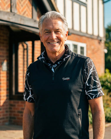 Man wearing a black Caddie Outfitters shirt standing in front of a brick building with greenery.