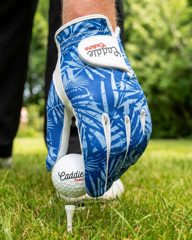 Golf glove with blue leaf pattern holding a golf ball on a tee, on a grassy background.