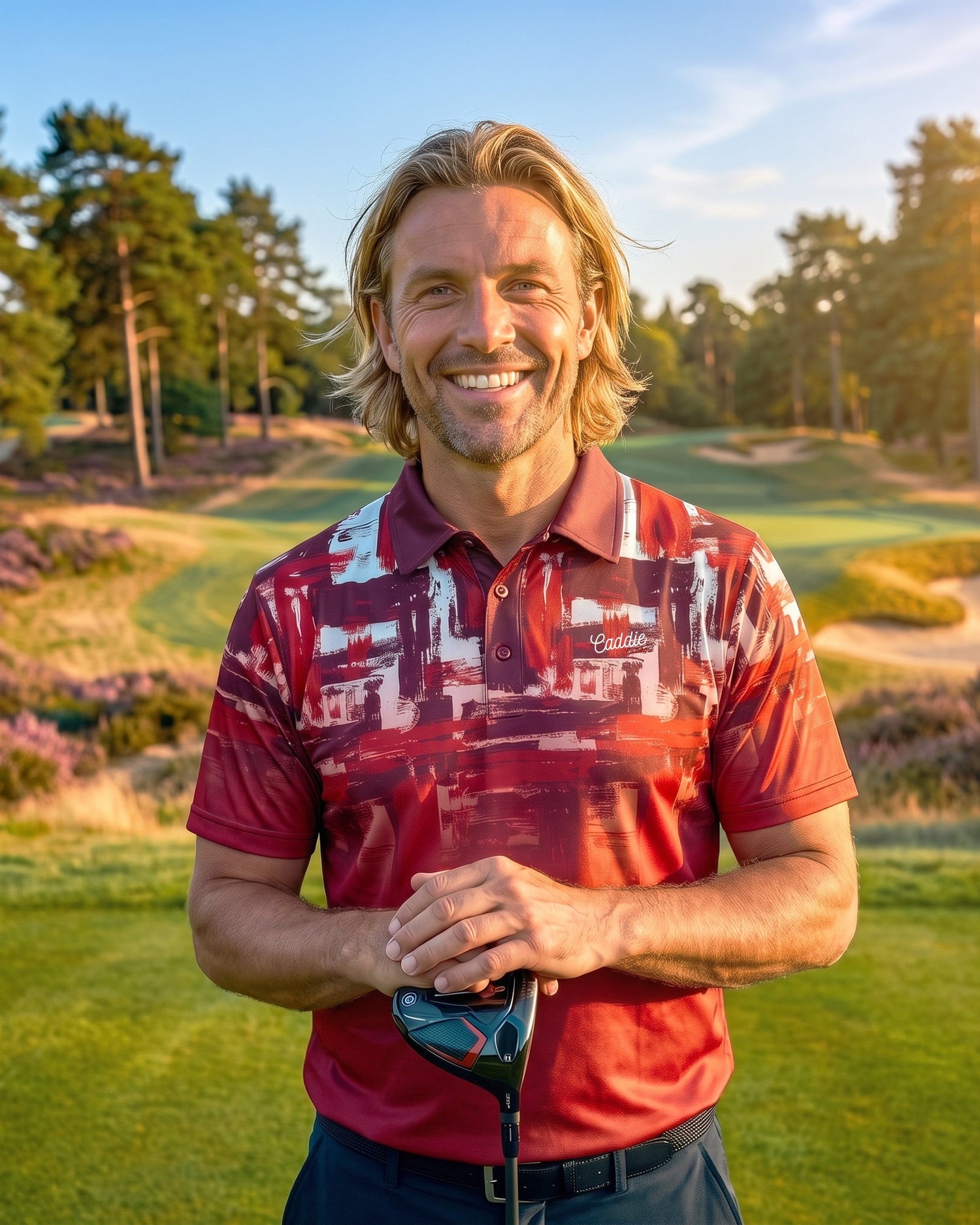 A smiling man with blonde hair stands on a golf course wearing a red and white patterned polo shirt, holding a golf club. Trees, grass, and sand bunkers are visible in the background during golden hour.
