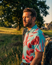 A man wearing a light blue short-sleeve polo shirt with a bold red tropical floral pattern, standing outdoors next to a stone wall in a grassy field with trees in the background.